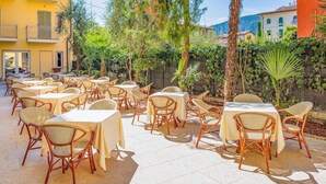 Une terrasse extérieure avec des tables et des chaises en osier, entourée de verdure et de bâtiments.