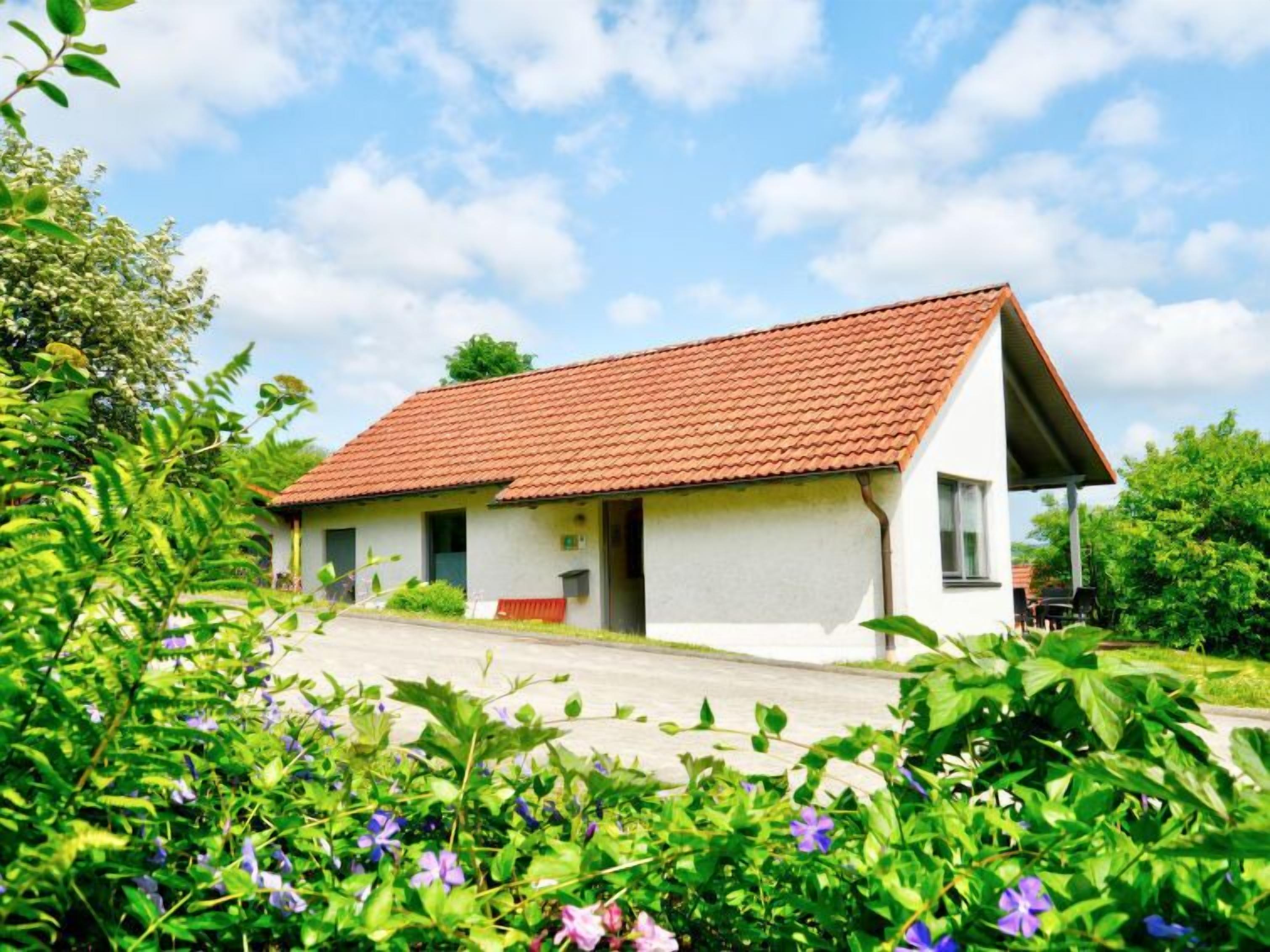 A single-story house with a red-tiled roof, a small porch, and a garden with various plants and flowers.
