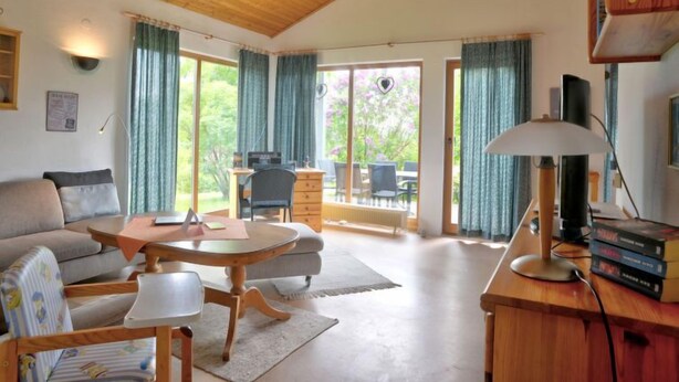 A spacious living room with a wooden ceiling, a sofa, a coffee table, a desk with a lamp, and a view of the outdoors through large windows. - Solling by Interhome (Uslar)