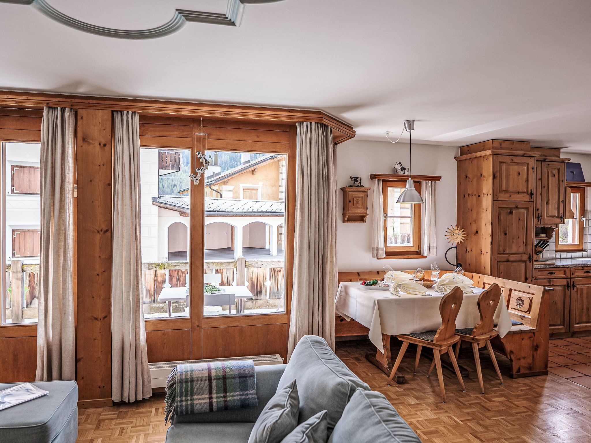 Ein gemütliches Wohnzimmer mit einer Couch, einem Esstisch mit Stühlen, Holzschränken und großen Fenstern mit Vorhängen.