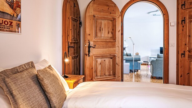 A bedroom with a bed, pillows, a nightstand, and a poster on the wall. There are wooden doors and a window leading to another room. - Chesa Mezzaun by Interhome (Zuoz)