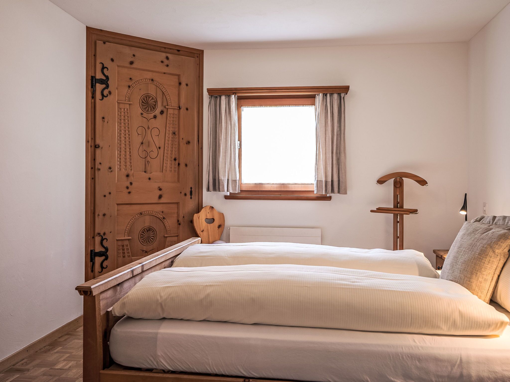 Ein Schlafzimmer mit Holzschrank, einem fenster mit Vorhängen, einem Nachttisch mit Lampe und einem Bett mit weißer Bettwäsche.