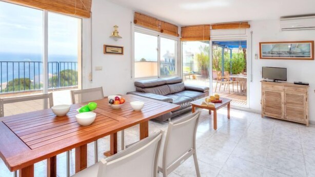 A spacious living area with a wooden dining table, a sofa, and a TV. Large windows offer a view of the sea.