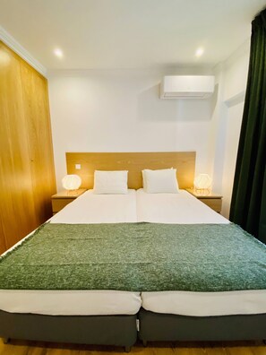 Ein Doppelbett mit grüner Decke, zwei Nachttische mit Lampen, ein Holz-Kopfteil und eine Decken-Klimaanlage.