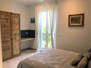 Een slaapkamer met een bed, een houten kledingkast, een raam met gordijnen, een klein plankje en een schilderij aan de muur.