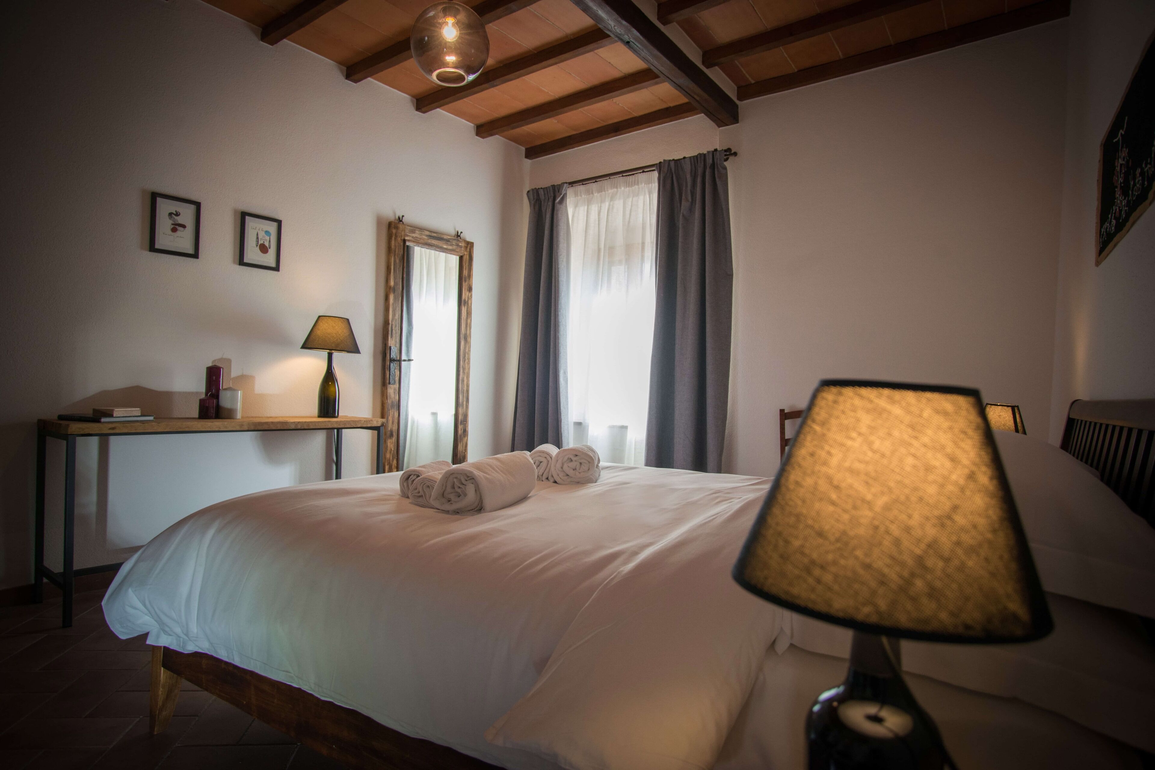 A neatly made bed with rolled towels, a bedside table with a lamp, a desk with a lamp, and a window with curtains.