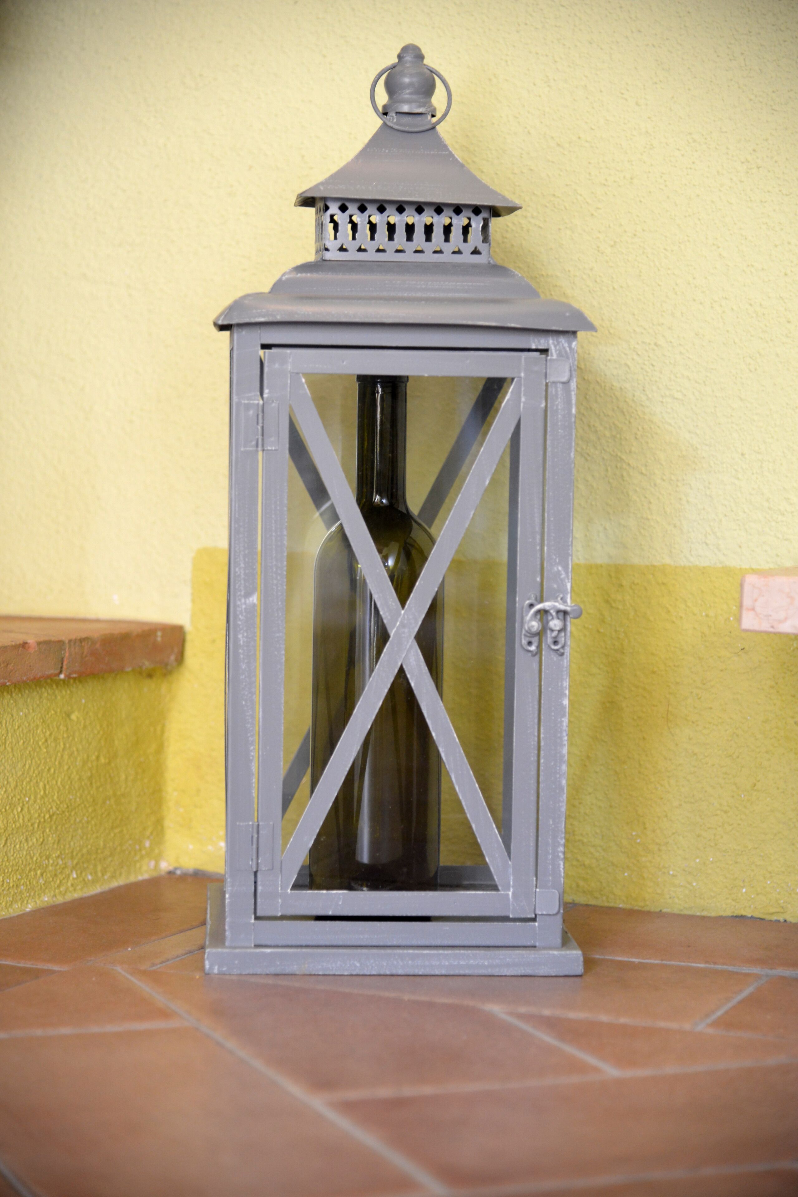 Une lanterne grise, dotée d’une porte en verre et d’une anse en métal, est posée sur un sol carrelé, adossée à un mur jaune.