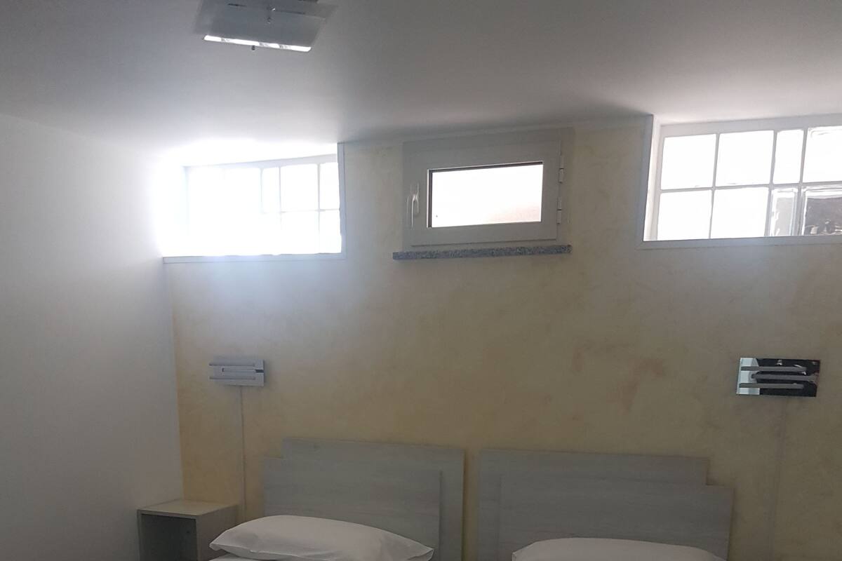 Una habitación con dos camas individuales, cada una con una almohada y una toalla doblada. Hay dos ventanas que dejan entrar luz natural y un equipo de aire acondicionado de pared.