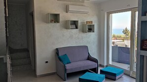 A modern living room with a grey sofa, teal cushions, and a glass door leading to a balcony with a view. - Alba De Mar (Roseto Capo Spulico)