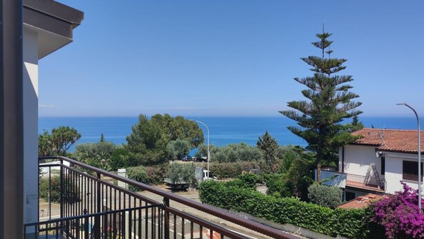 A balcony with a view of the sea, surrounded by greenery and a building with a tiled roof. - Alba De Mar (Roseto Capo Spulico)