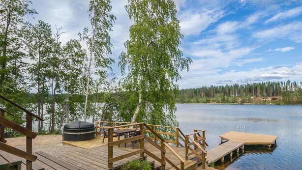 Una terraza de madera con un jacuzzi, mesas de picnic y un muelle que se extiende sobre un lago, rodeado de árboles.