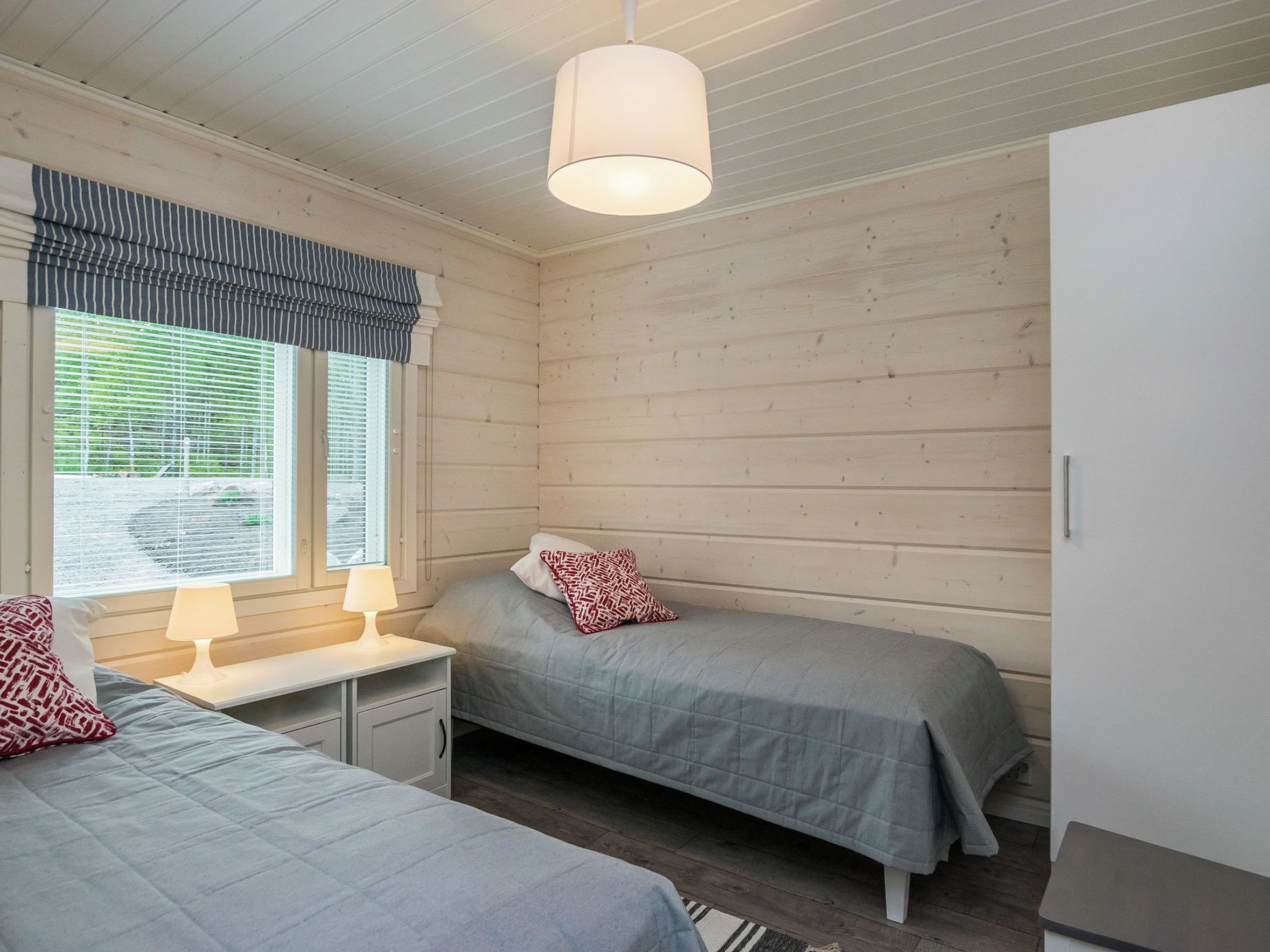 A room with two beds, a window with blinds, a lamp, and a wardrobe.