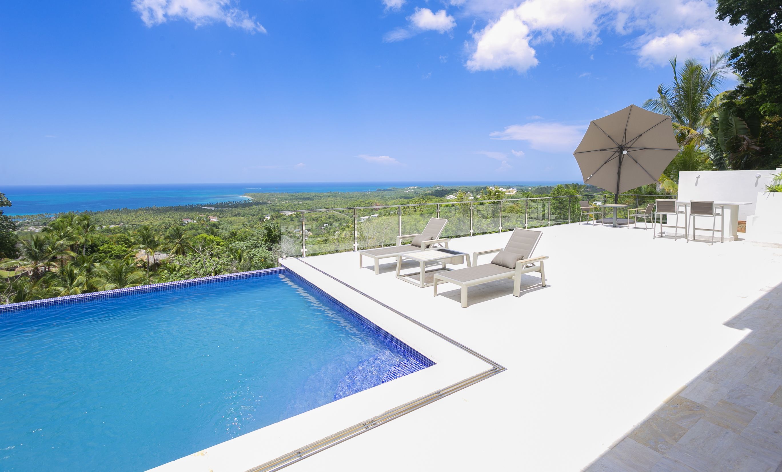 Piscina infinita en la azotea con sillones de descanso y una sombrilla, con vistas a un paisaje costero.