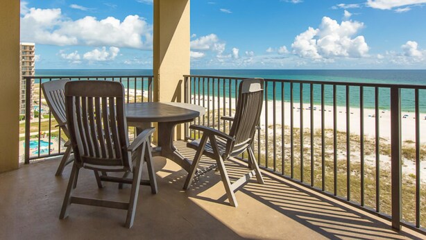 Un balcon avec deux chaises et une table, donnant sur une plage d’où l’on a une vue dégagée sur l’océan et un bâtiment situé au loin.