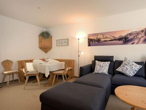 A cozy living room with a wooden dining table set for two, a dark sofa with patterned cushions, and a large painting of a mountainous landscape on the wall. - Chesa Flora 53 by Interhome (St. Moritz)
