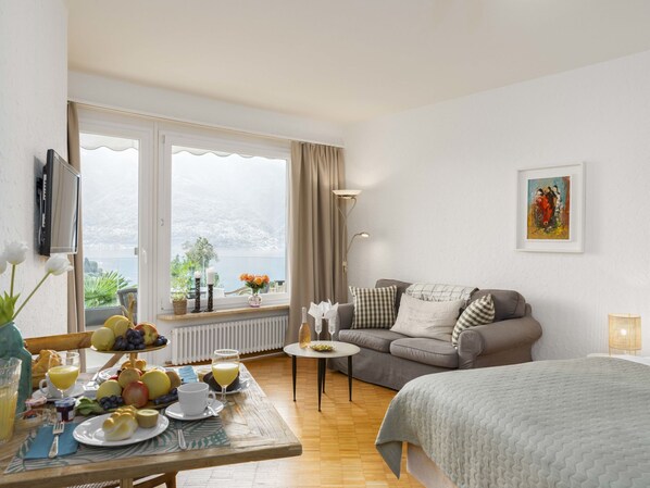 A hotel room with a bed, a sofa, a dining table with breakfast, a TV, and a large window with a view of the sea.