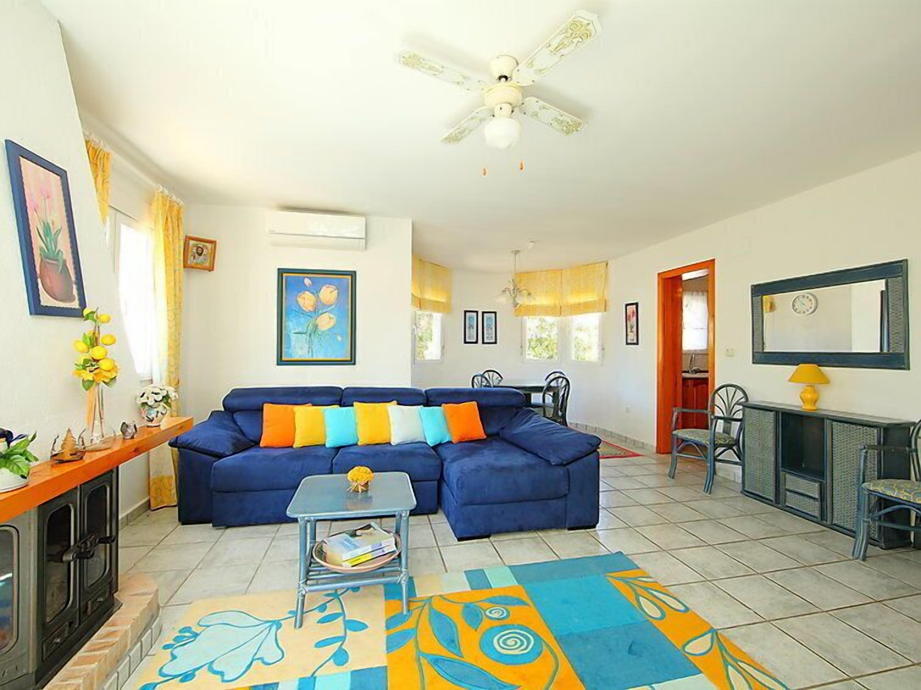 A living room with a blue sofa, a small table, a ceiling fan, and a colorful rug.