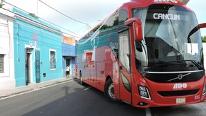 En röd buss med texten ”CANCUN” på framsidan står parkerad på en gata, och i bakgrunden syns en blå byggnad med skylten ”GUAYA HOSTEL”.