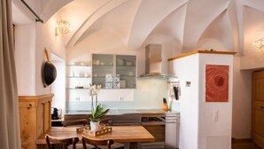 A modern kitchen with a wooden dining table, chairs, and a central island. There is a window with shelves above the counter, and a red abstract painting on the wall. - Chesa Chascharia 1 by Interhome (Samedan)