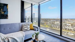 A modern living room with a grey sofa, a small round table, and a large window offering a city view.
