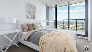 A modern bedroom with a large bed, a bedside table, a nightstand, a window with a city view, and a wall-mounted artwork.