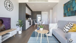 A modern living room with a grey sofa, a coffee table, and a TV. There is a kitchen in the background with a dining area.