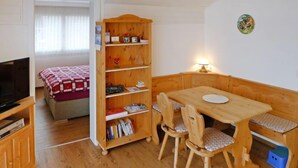 A cozy room with a wooden dining table, two chairs, a bookshelf, a television, and a bed with a red and white checkered blanket. - Apartment Ladasa by Interhome (Emmetten)