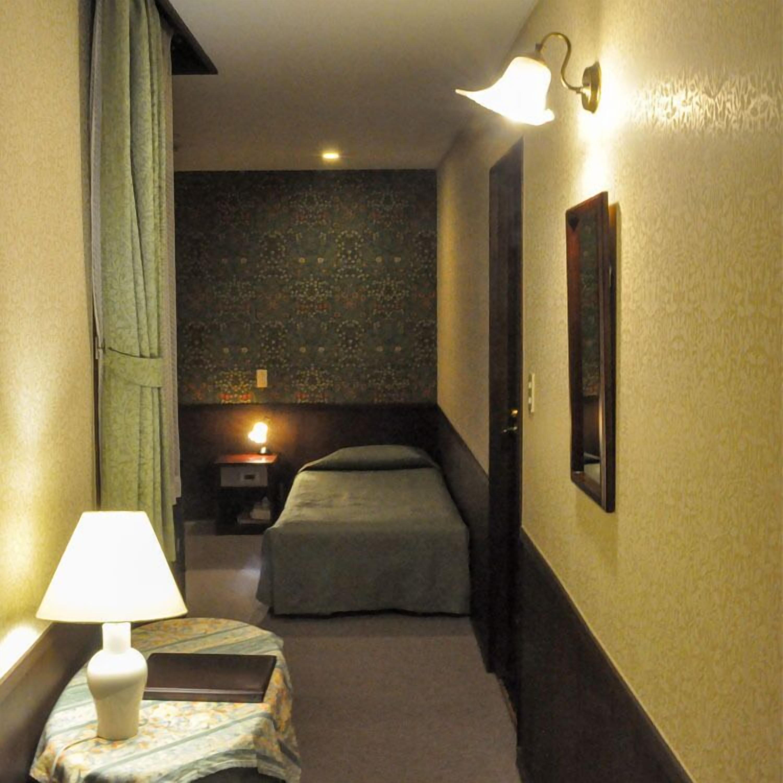 A narrow hallway with a bed, a nightstand, a lamp, and a wall-mounted light fixture.