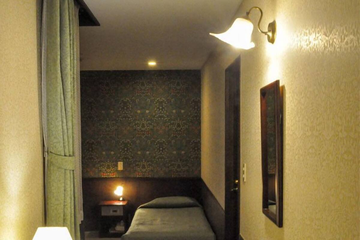A narrow hallway with a bed, a nightstand, a lamp, and a wall-mounted light fixture.