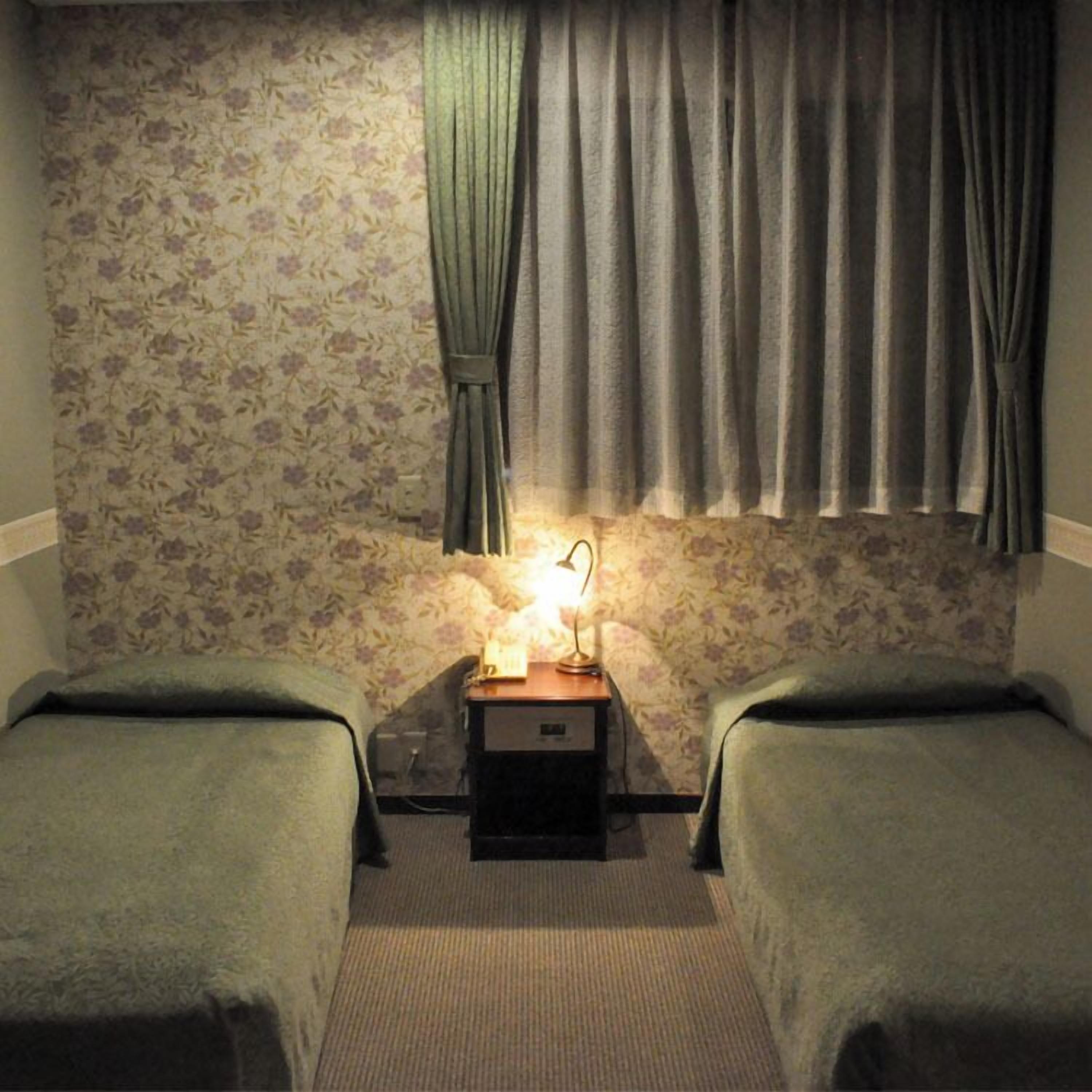 Una habitación con dos camas individuales, una mesita de noche con lámpara, papel tapiz con motivos florales y una ventana con cortinas.