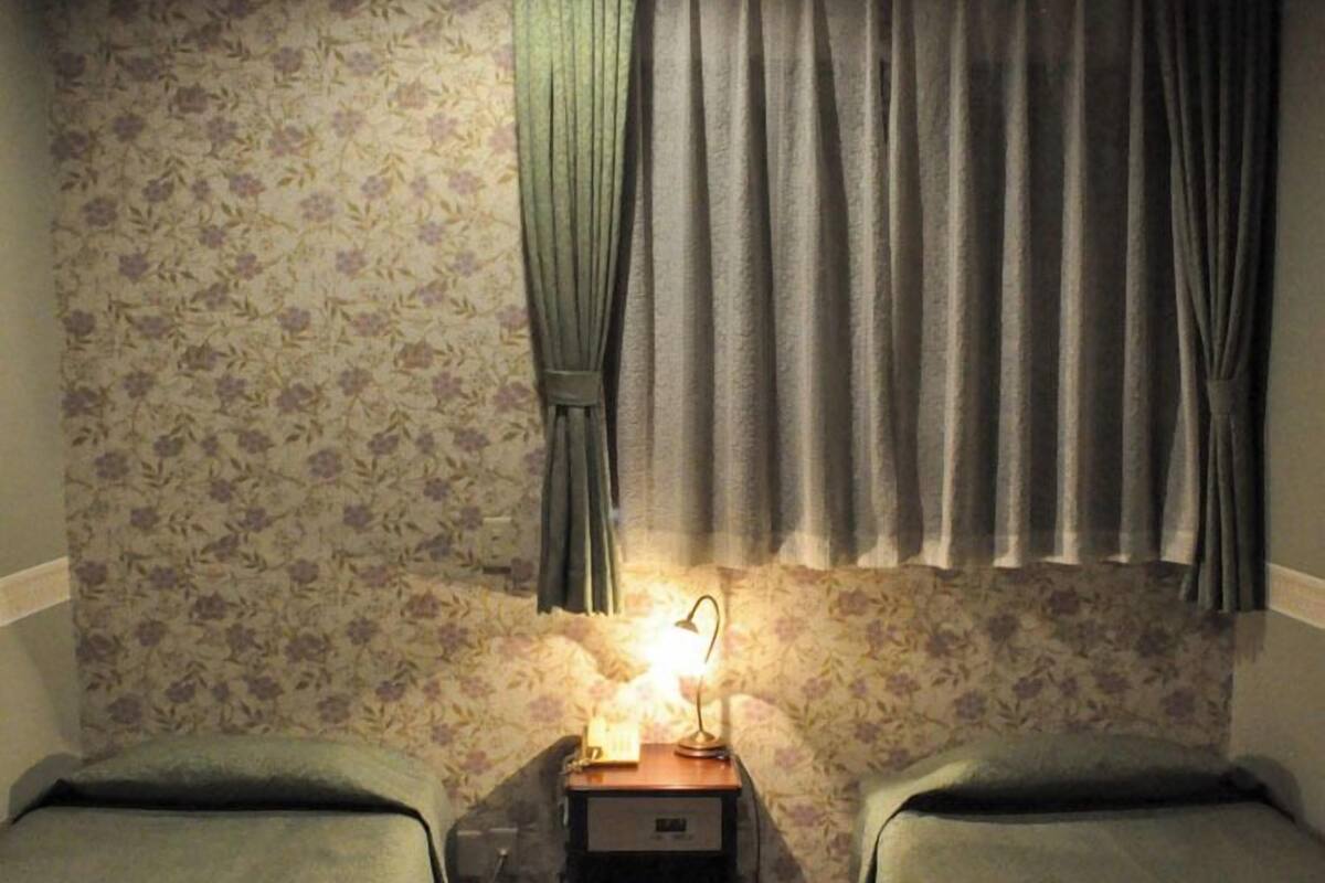 A room with two single beds, a nightstand with a lamp, floral wallpaper, and a window with curtains.