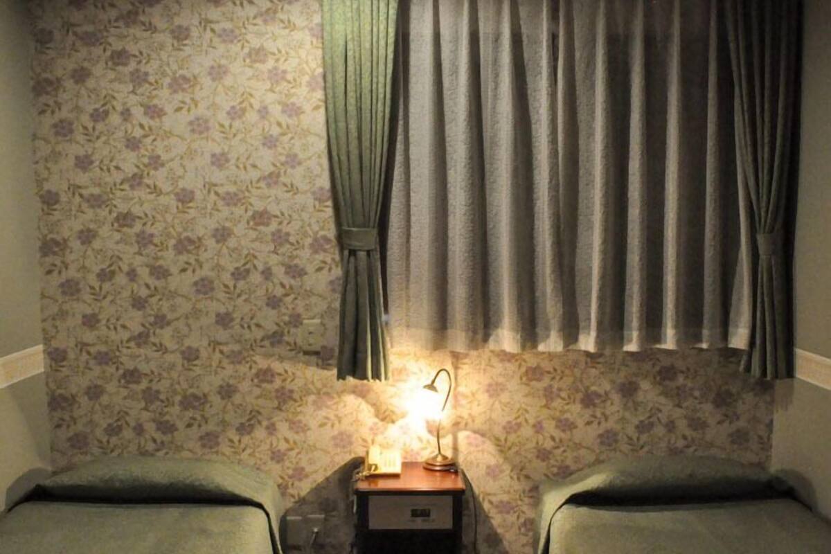 A room with two beds, a nightstand with a lamp, floral wallpaper, and curtains.