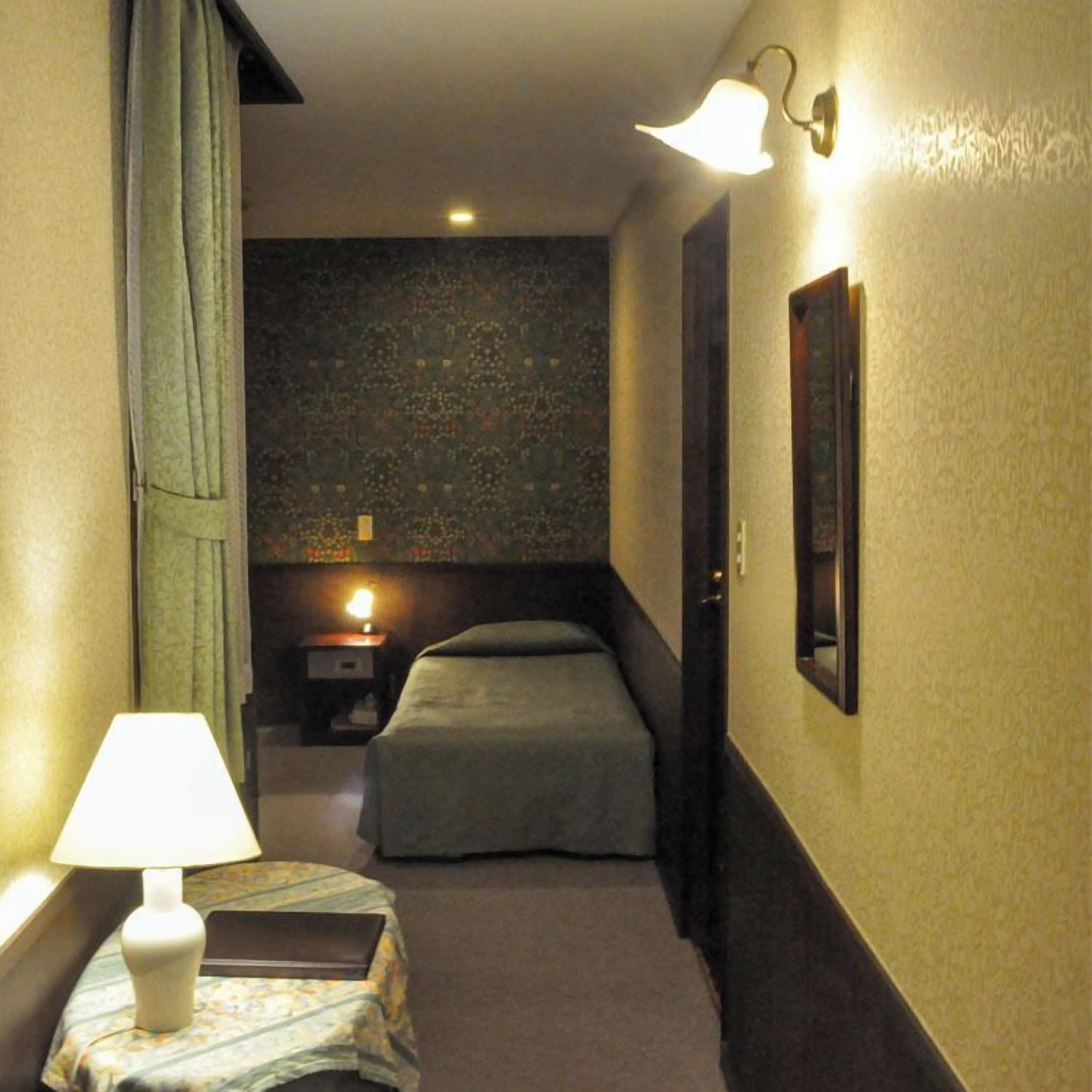 Un pasillo estrecho con una cama, una mesita de noche, una lámpara y un aplique de pared.