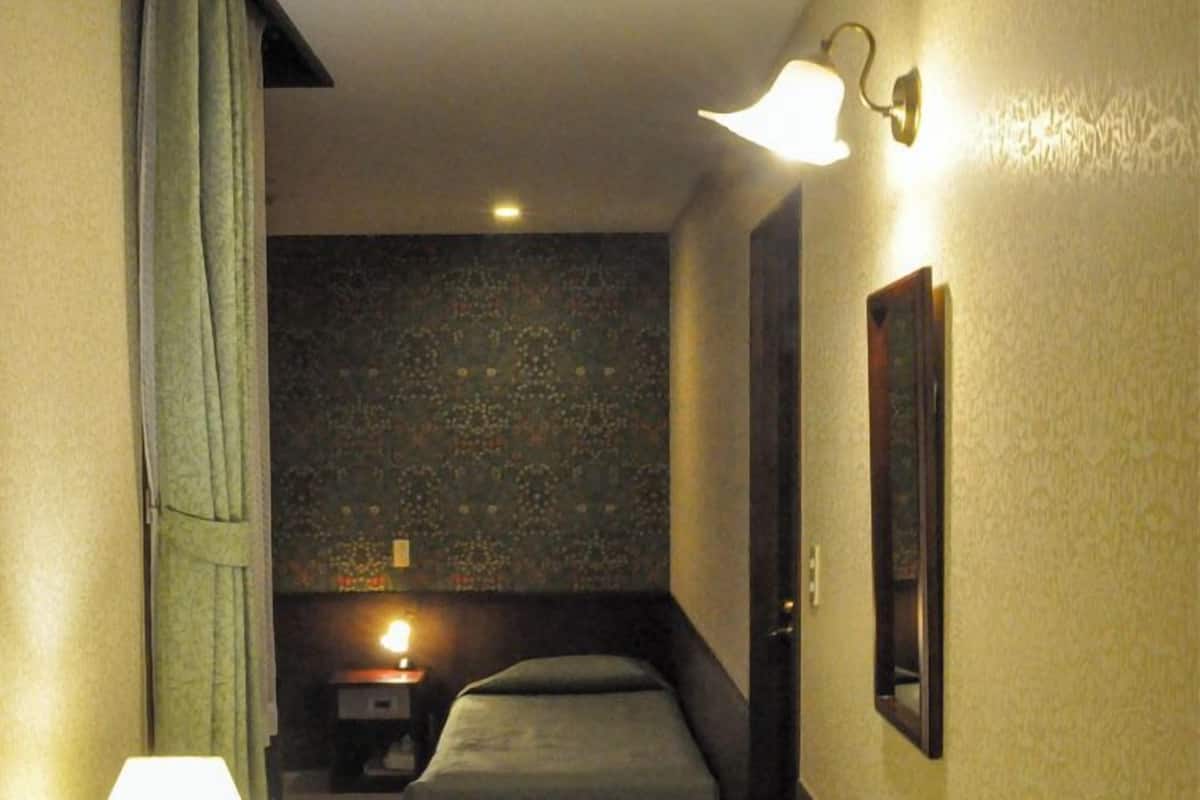 Un couloir étroit comprenant un lit, une table de chevet, une lampe et un luminaire fixé au mur.