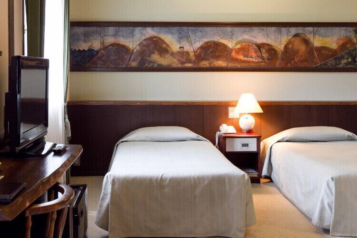 A hotel room with two beds, a television, a painting on the wall, and a lamp on the nightstand.