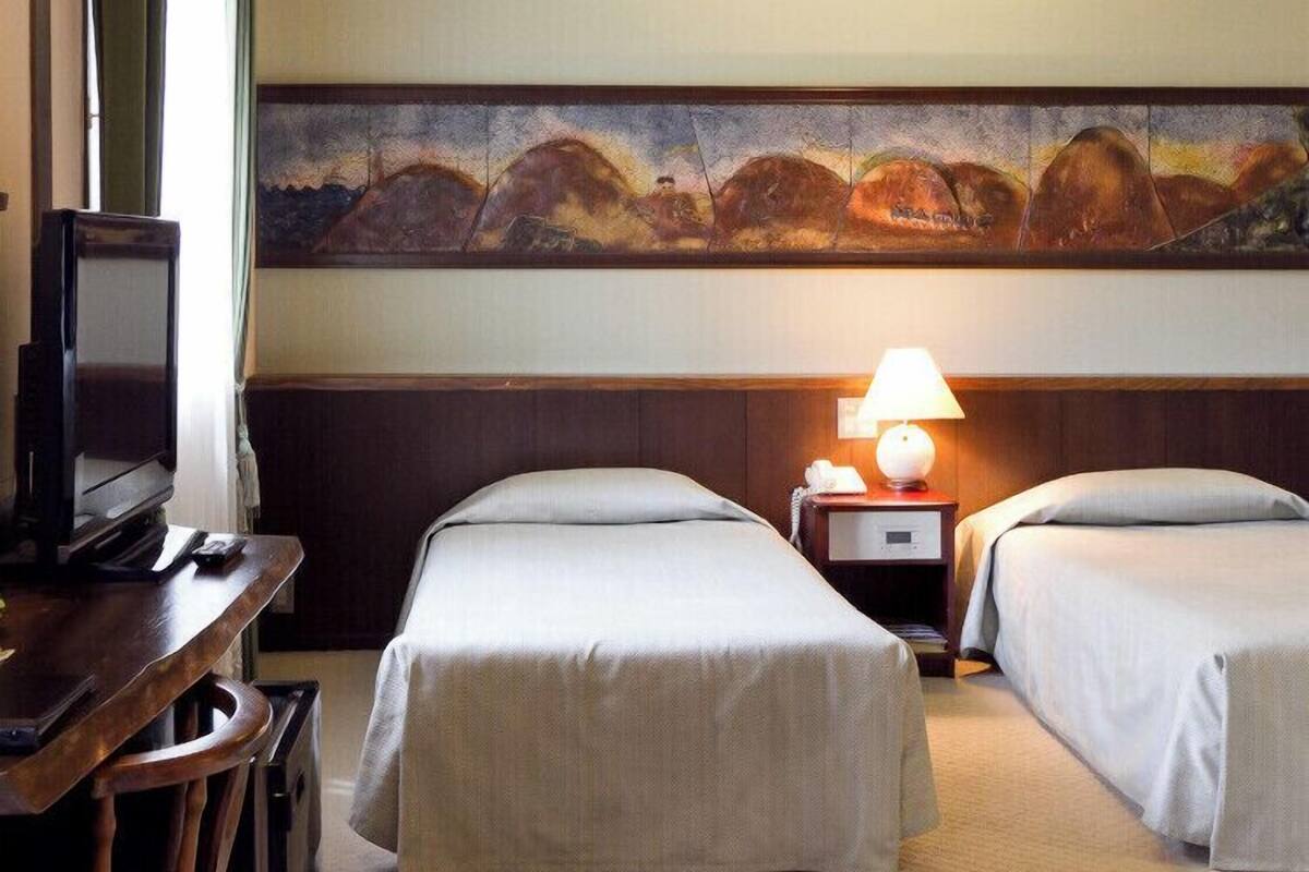 A hotel room with two beds, a television, a painting on the wall, and a lamp on the nightstand.