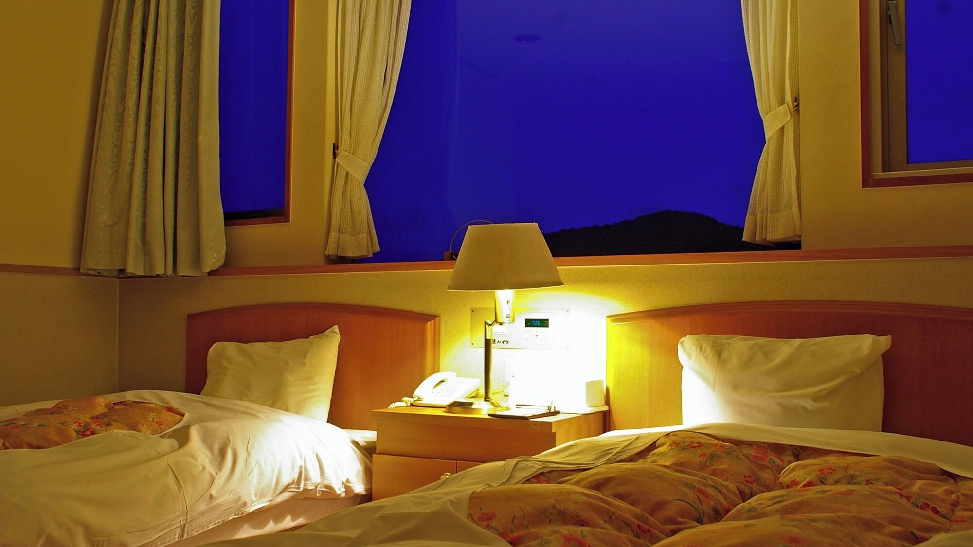 Habitación de hotel con dos camas, una mesita de noche con lámpara y una ventana con cortinas.