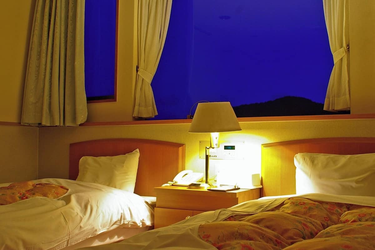 Habitación de hotel con dos camas, una mesita de noche con lámpara y una ventana con cortinas.