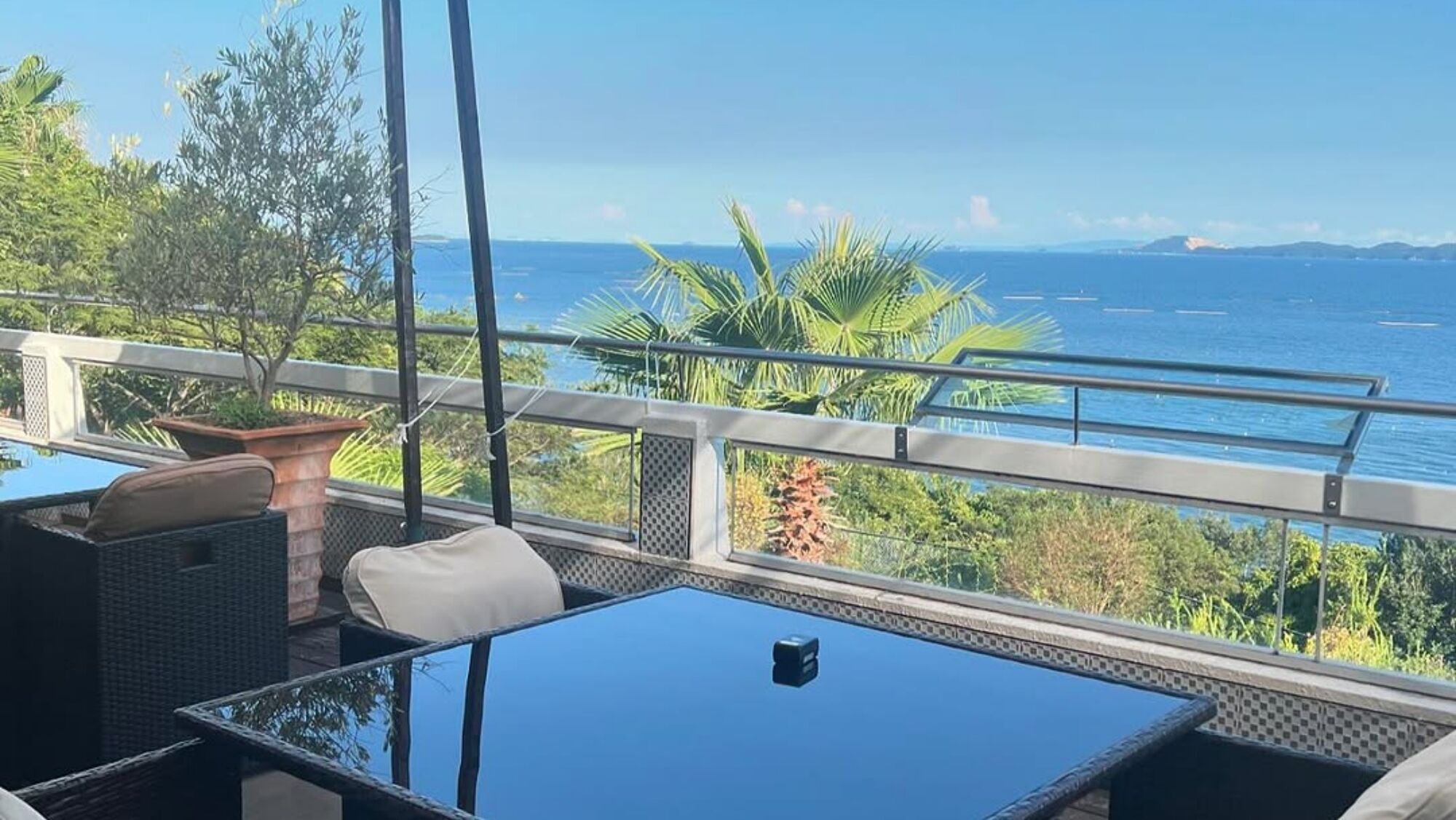 A balcony with a glass table, wicker furniture, and a view of the sea and distant mountains.