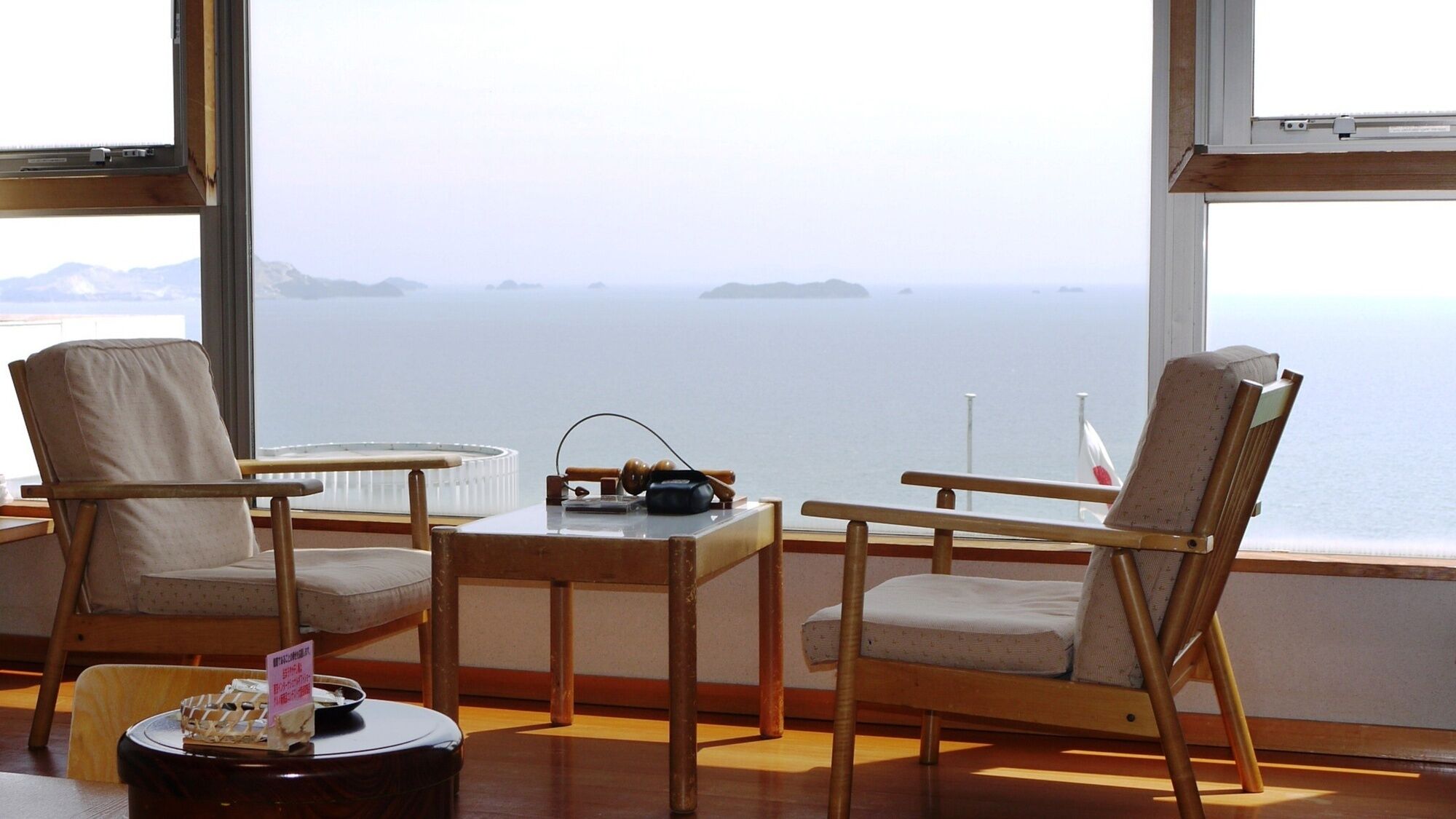 A room with two wooden chairs, a small table, and a view of the sea through large windows.