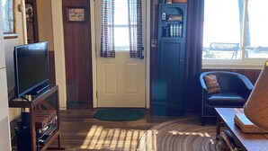 Una sala de estar con un televisor sobre un mueble de madera, una puerta con cortinas, una estantería, un sillón de cuero oscuro y una alfombra tejida.