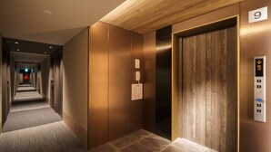 A modern elevator with a digital display, a wooden panel design, and a carpeted hallway leading to it. - Super Hotel Anan Tomioka (Anan)