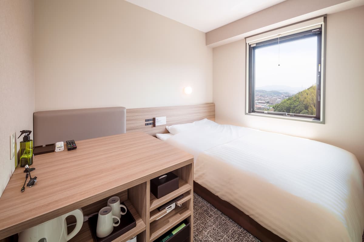Hotelkamer met een bed, een bureau met stoel, een raam met uitzicht op de stad en een nachtkastje met een lamp en een fles.