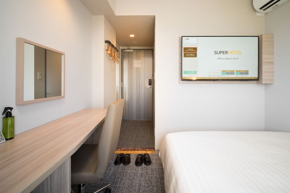 Een hotelkamer met een bed, een bureau, een stoel, een aan de muur bevestigde televisie en een schoenenrek.