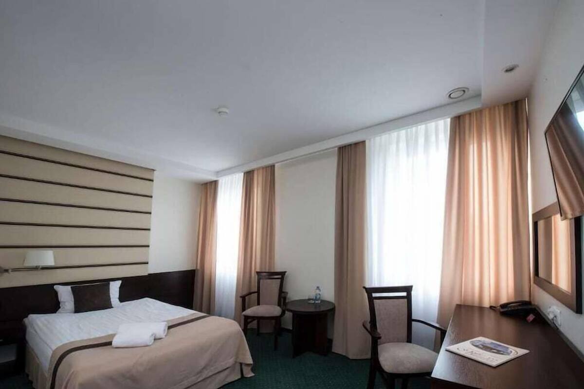 Hotellihuone, jossa on sänky, kaksi tuolia, pieni pöytä, televisio ja suuret ikkunat, joissa on verhot.