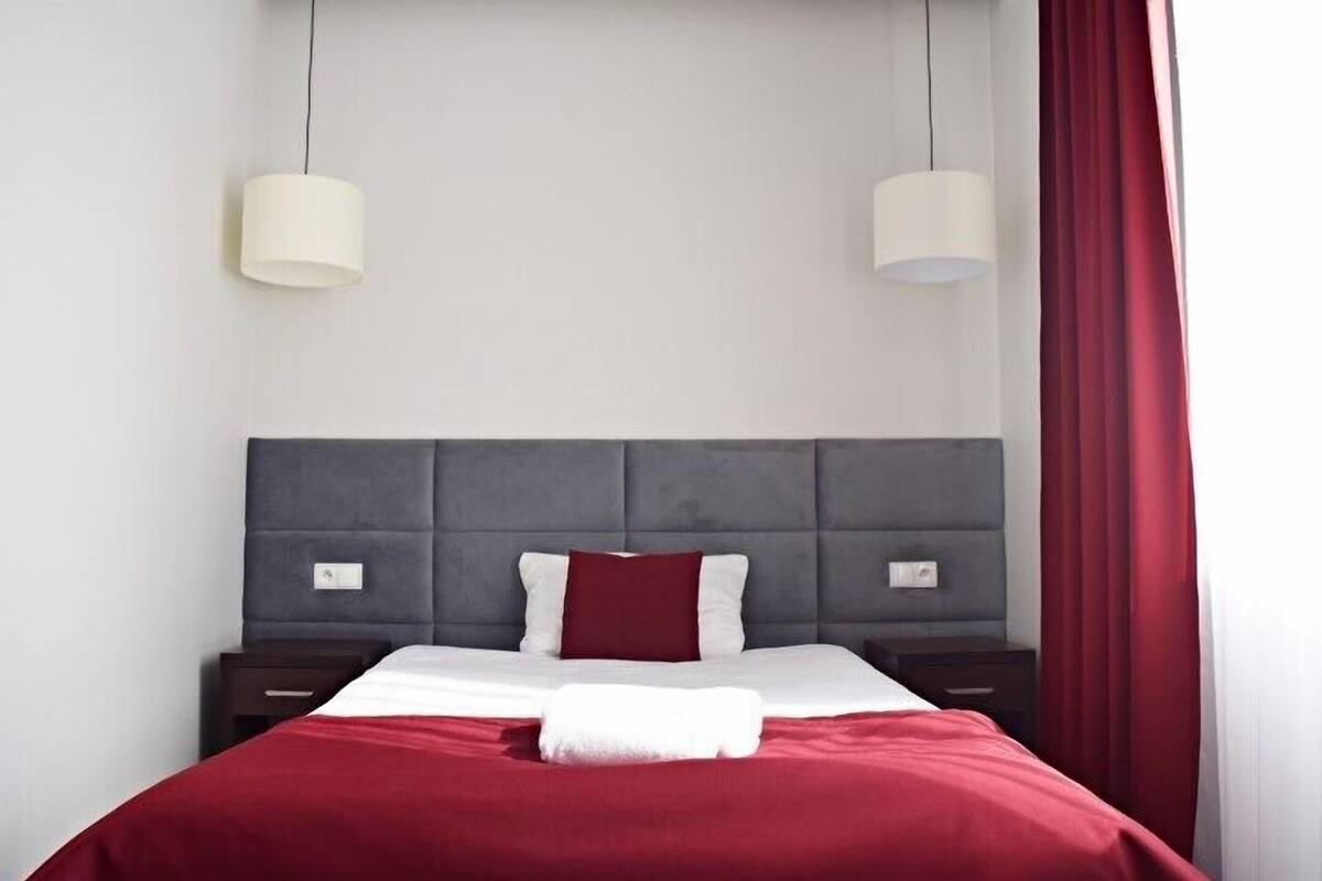 Hotellihuone, jossa on sänky, kaksi yöpöytää, punainen verho ja kaksi riippuvalaisinta.