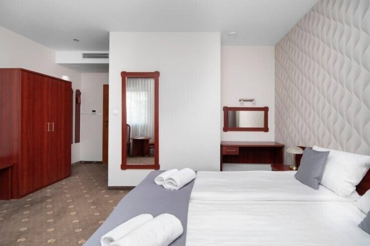 Hotellihuone, jossa on suuri sänky, puinen vaatekaappi, peili, yöpöytä ja pieni työpöytä, jossa on lamppu.