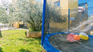 A trampoline with a safety net, surrounded by a garden with a tree and a house in the background.