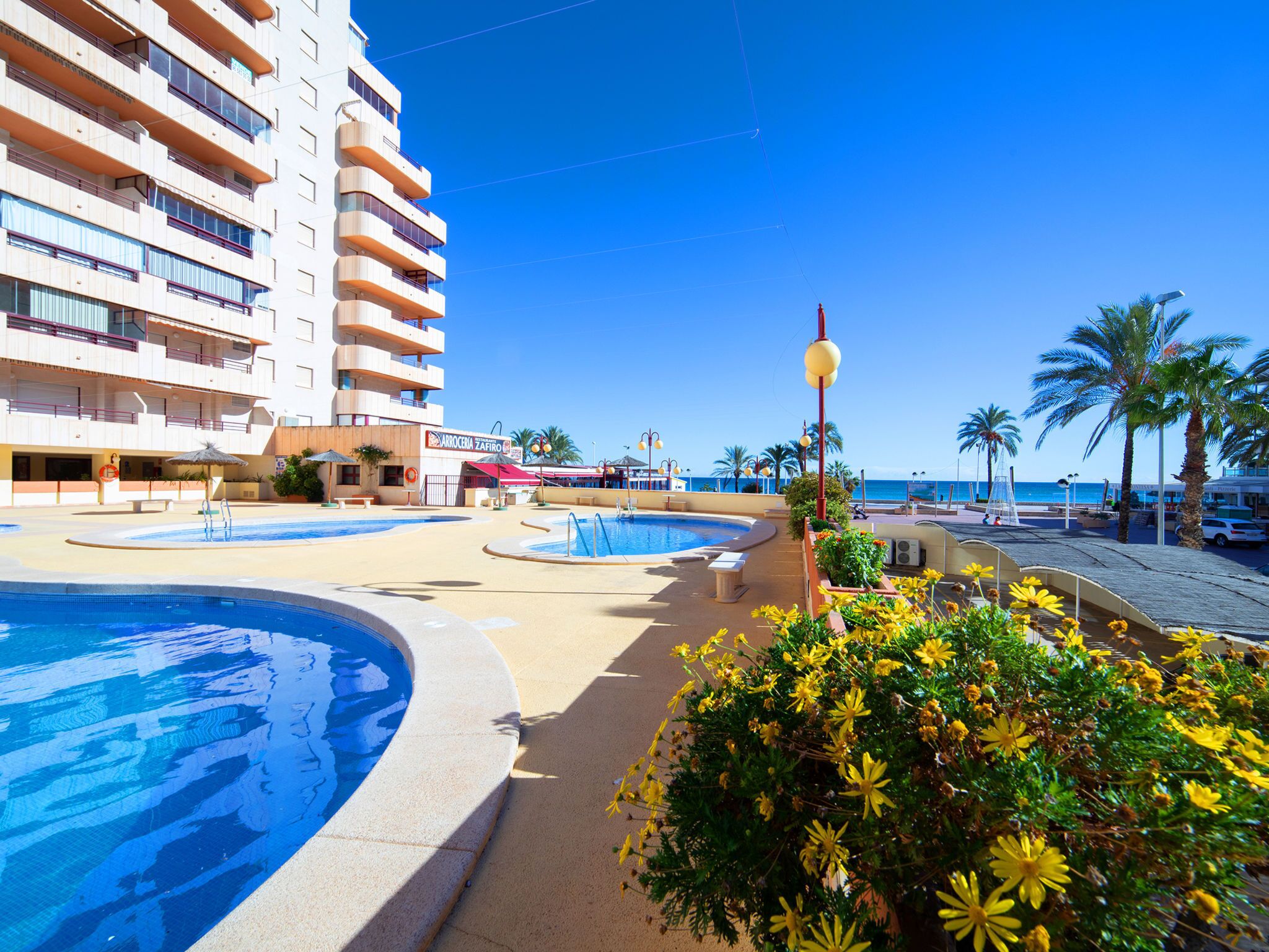 A resort pool area with multiple swimming pools, a building with balconies, and a clear view of the sea.