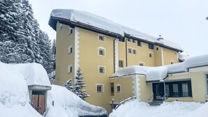 Un immeuble à plusieurs étages, doté d’une façade jaune, entouré d’arbres couverts de neige et surmonté d’un toit également enneigé.